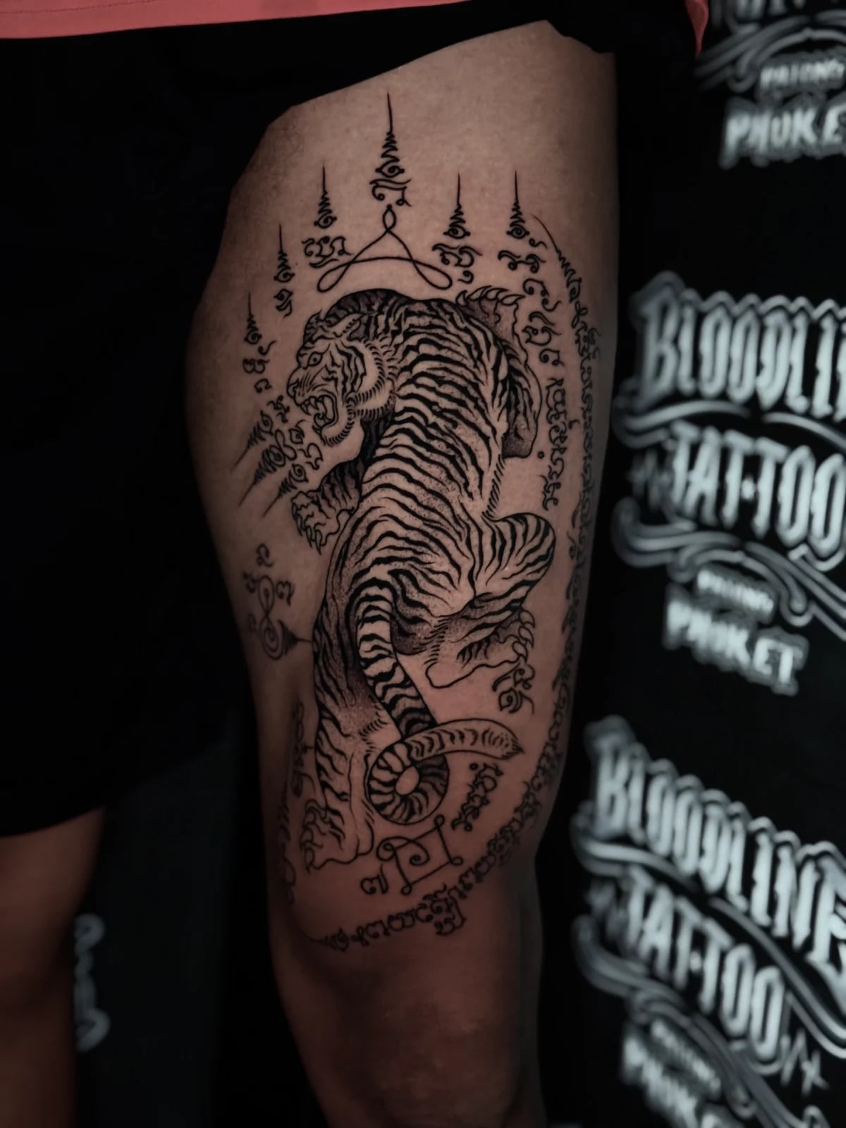 Suea (Tiger) Sak Yant of a snarling tiger with Khom script border and multiple unalomes on the outer thigh at Bloodline Tattoo Phuket.