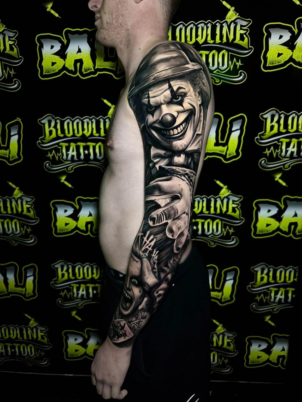 Detailed black and grey Chicano full arm sleeve tattoo with a grinning clown in a bowler hat, dramatic pointing hand, and screaming face below at Bloodline Tattoo Bali.