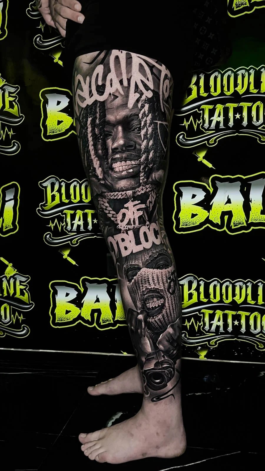 Detailed black and grey Chicano thigh and shin tattoo with a braided rapper portrait, bold script, ski mask figure, and handgun at Bloodline Tattoo Bali.