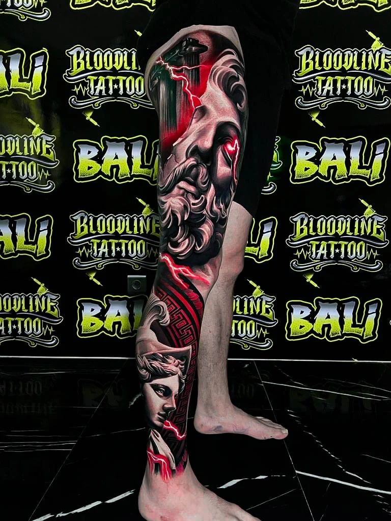 Colour mythology leg sleeve tattoo with a bearded stone god, red lightning, and a dramatic female portrait below at Bloodline Tattoo Bali.