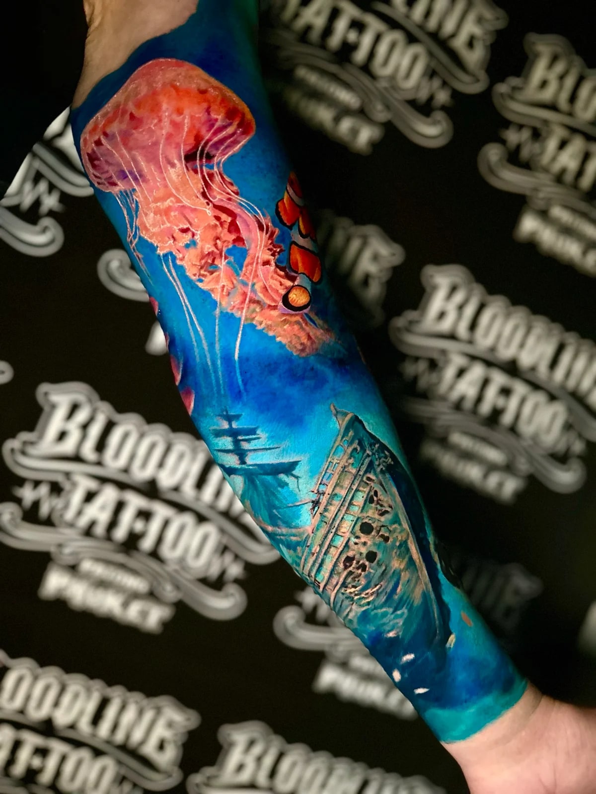 Underwater colour sleeve with a pink jellyfish, a clownfish, and a detailed sunken shipwreck in saturated blue ocean tones at Bloodline Tattoo Phuket.