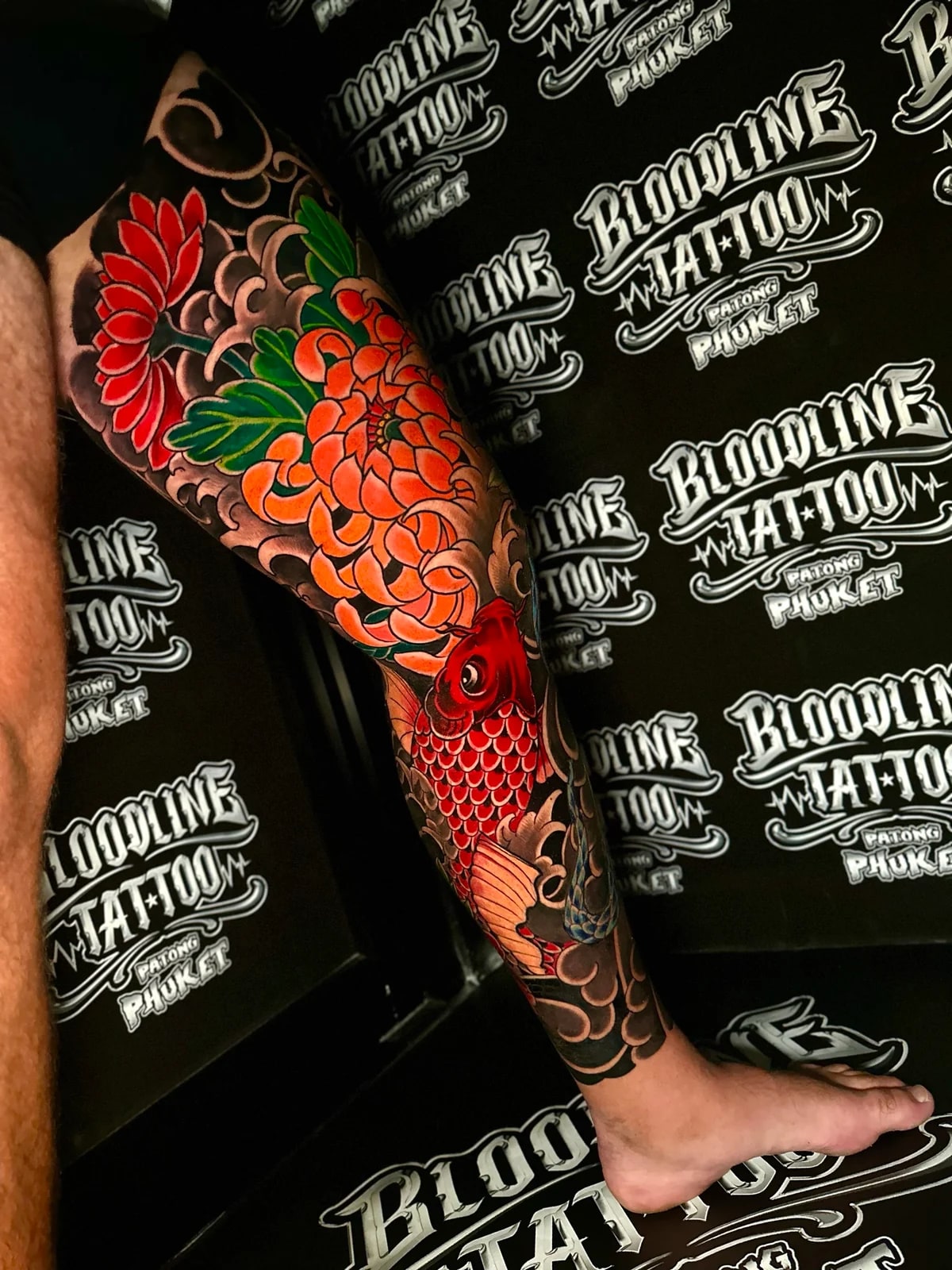 Japanese colour leg tattoo with a large orange chrysanthemum, a descending red koi fish, and dark swirling cloud backgrounds at Bloodline Tattoo Phuket.