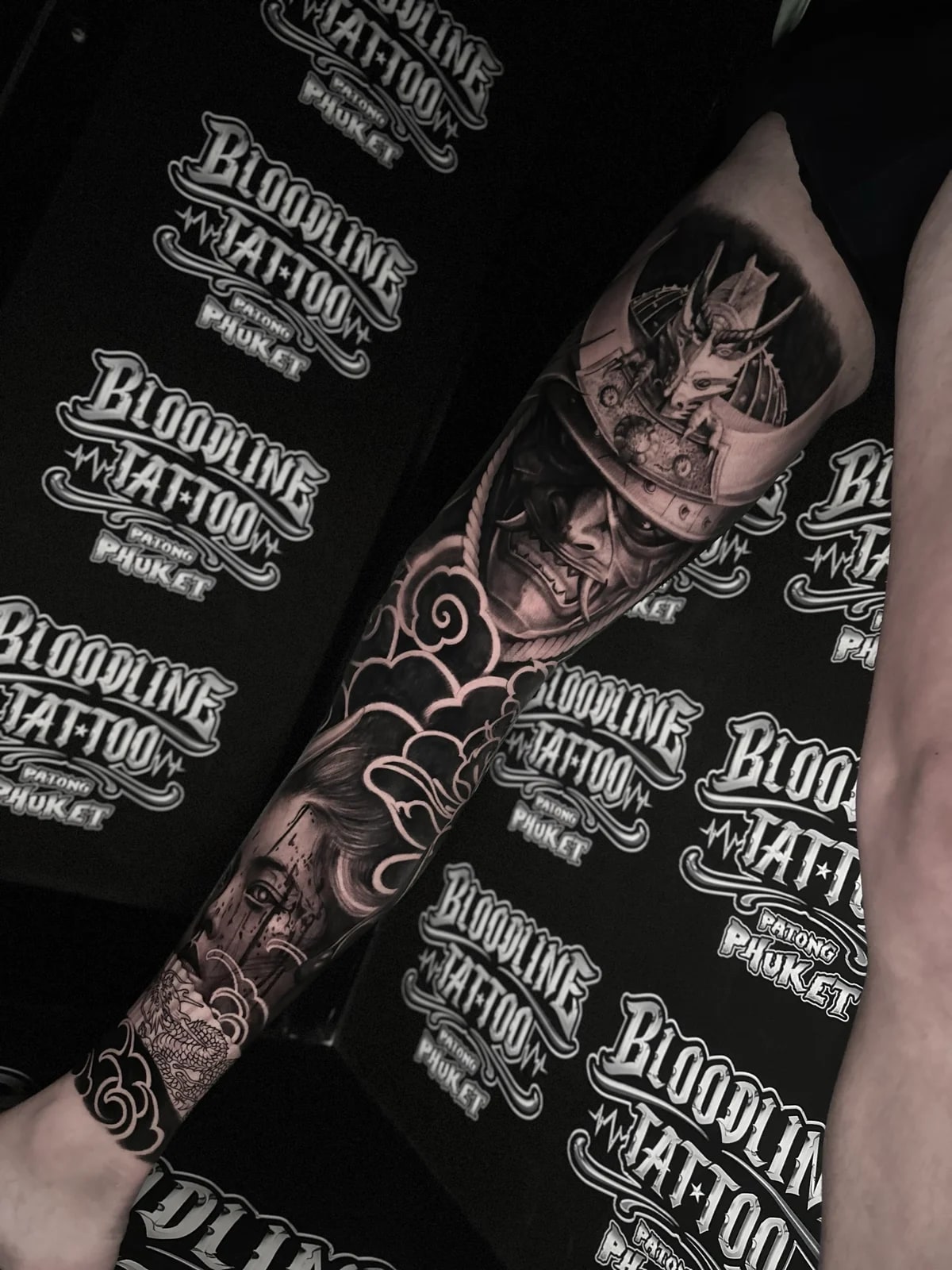 Japanese black and grey leg sleeve with samurai kabuto and menpo, woman's portrait on the calf, and dragon scales at the ankle amid clouds at Bloodline Tattoo Phuket.