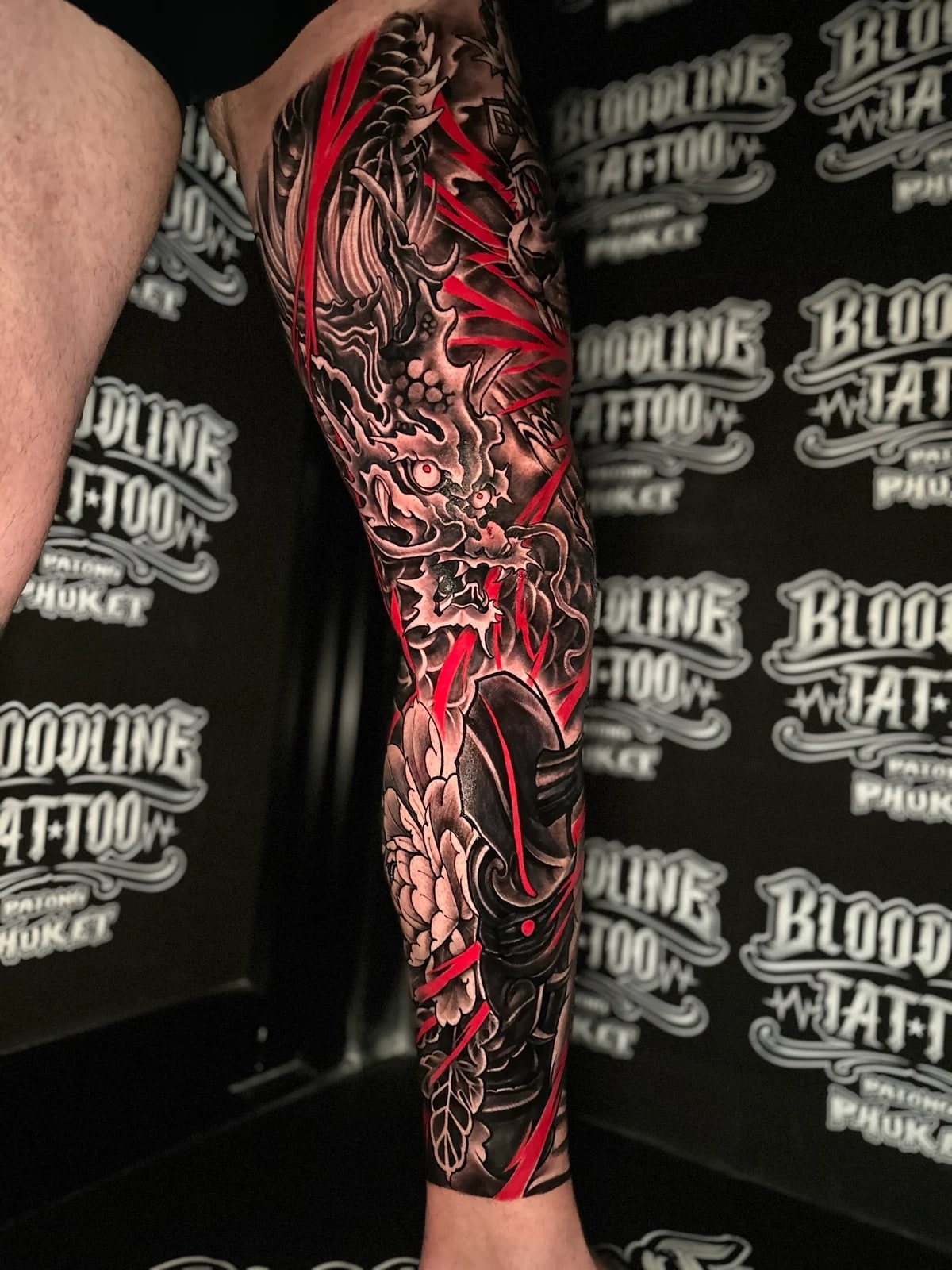 Japanese black and grey leg sleeve with dragon over the knee, peony on the shin, red lightning streaks, and shrine detail amid wind bars at Bloodline Tattoo Phuket.