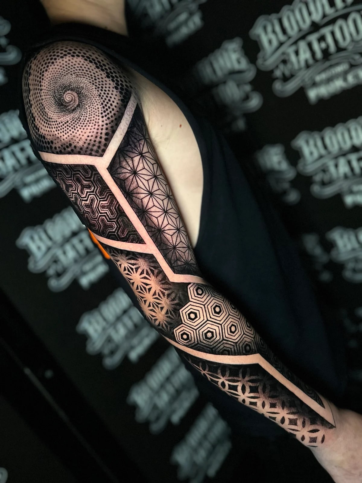 Panelled geometric mandala sleeve with a dotwork spiral shoulder, hexagonal grids, Flower of Life sections, and honeycomb forearm fill at Bloodline Tattoo Phuket.
