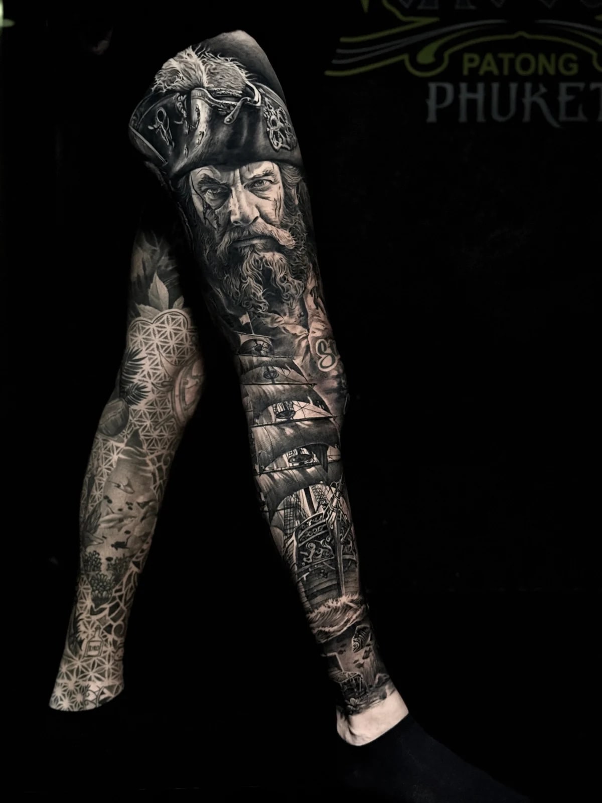 Detailed black and grey realism full leg sleeve tattoo featuring a pirate portrait, sailing galleon on waves, and underwater treasure chest at Bloodline Tattoo Phuket.