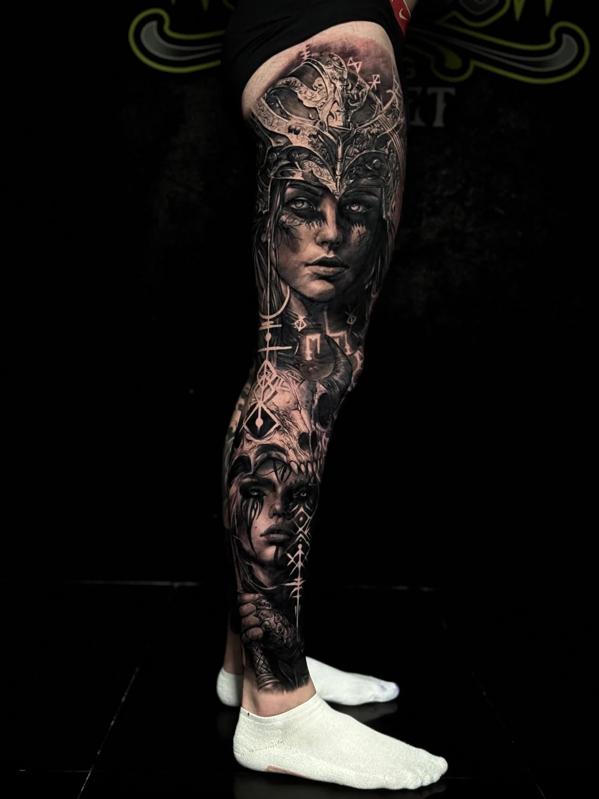 Detailed black and grey realism full leg sleeve tattoo featuring a Viking woman in an ornate helmet, ram skull, tear-streaked portrait, and sword hilt at Bloodline Tattoo Phuket.