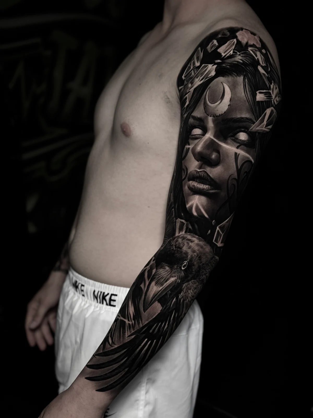 Detailed black and grey realism full sleeve tattoo featuring a mystical woman with crescent moon and crystal shards, and a detailed raven on the forearm at Bloodline Tattoo Phuket.