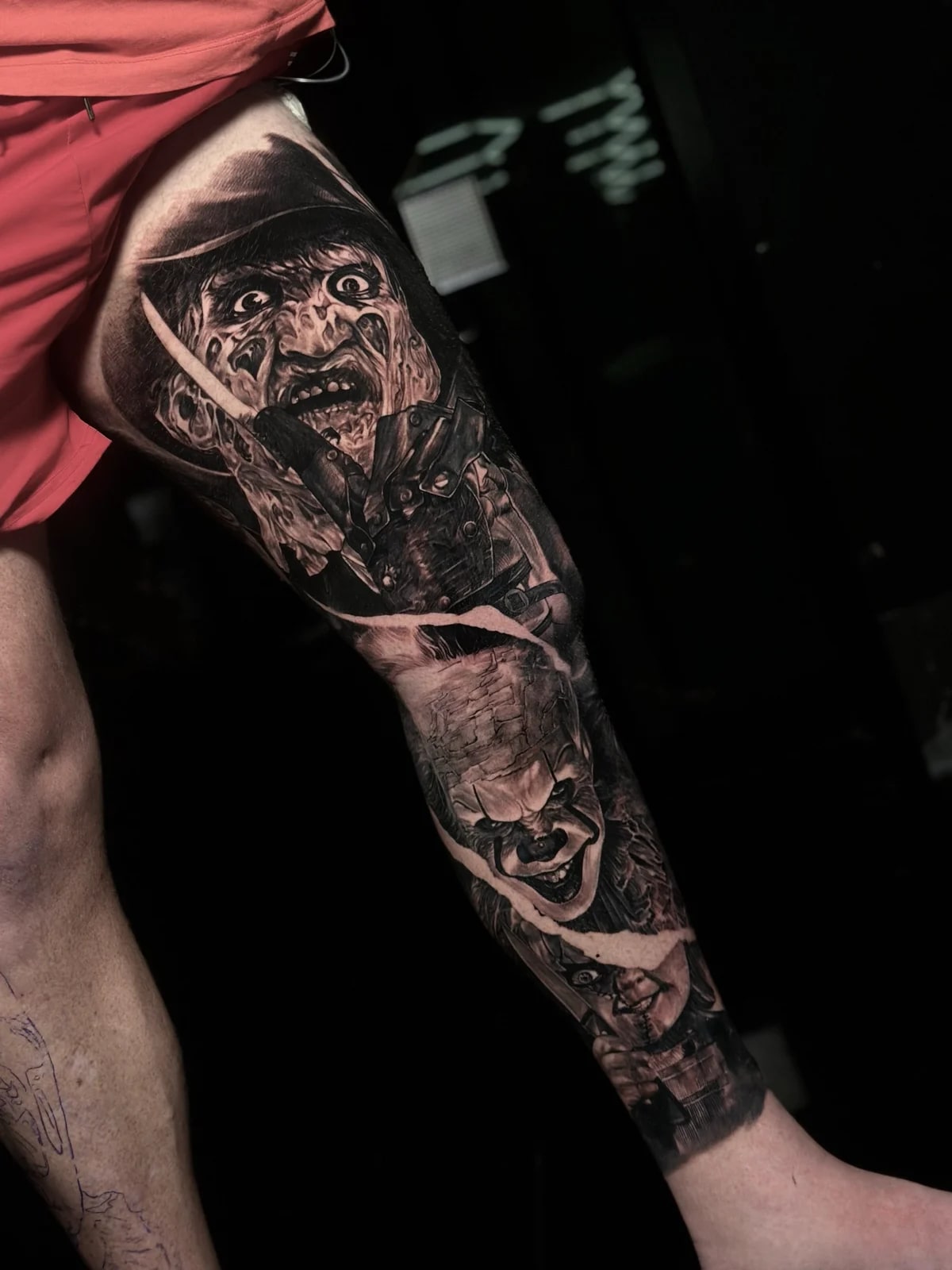 Detailed black and grey realism full leg sleeve tattoo featuring Freddy Krueger, Pennywise, and Chucky with a torn-paper effect at Bloodline Tattoo Phuket.